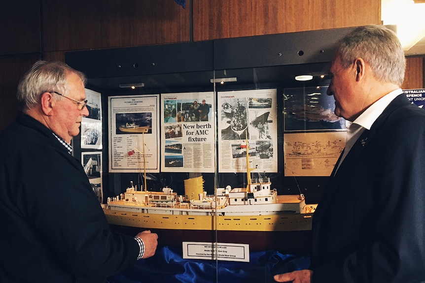 Two men look at a model ship behind glass.