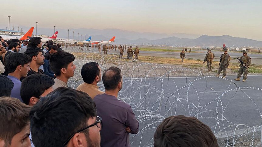 Afghan civilians behind barbed wire are watched by US Military who are inside the airport and planes are behind them.