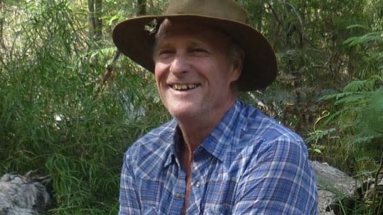 A man wearing a broad-brimmed hat, shorts and a checked shirt sits smiling in the bush.