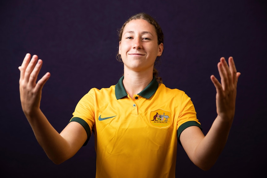 A female soccer player wearing yellow and green holds her arms wide