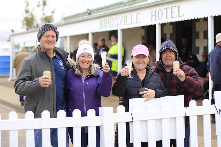 four people stand outside an old pub and behind a white fence with glasses in hand