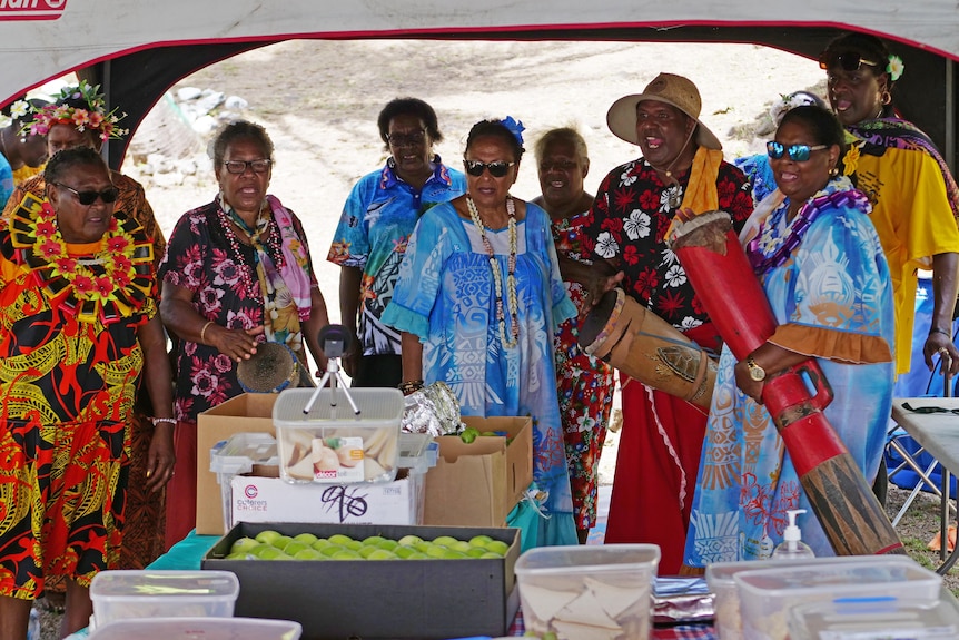 Group of people stand around a table of food singing.