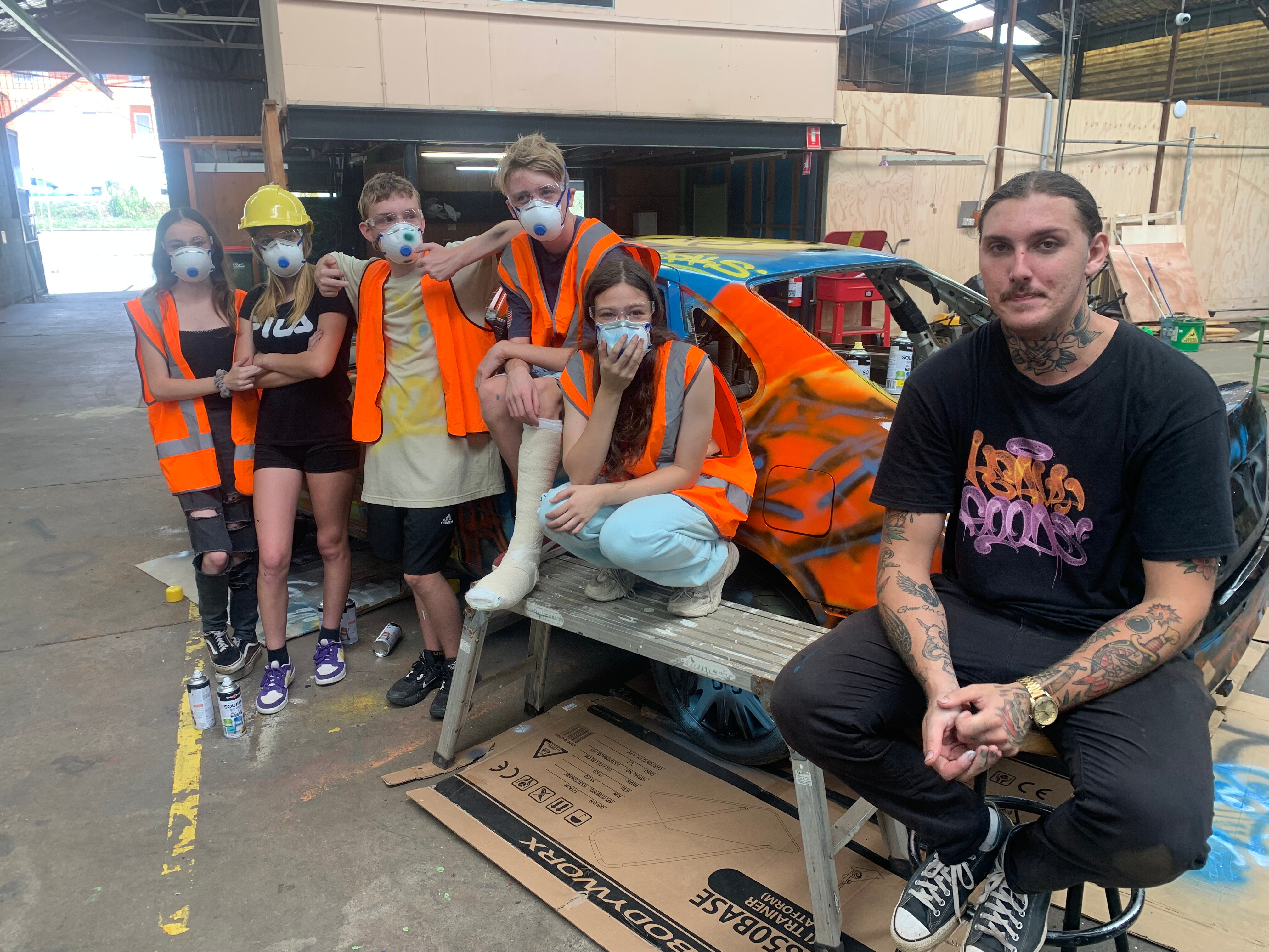 Five students in masks and hi-vis vests and a young man sit in front of a colourful spray-painted car in a shed.