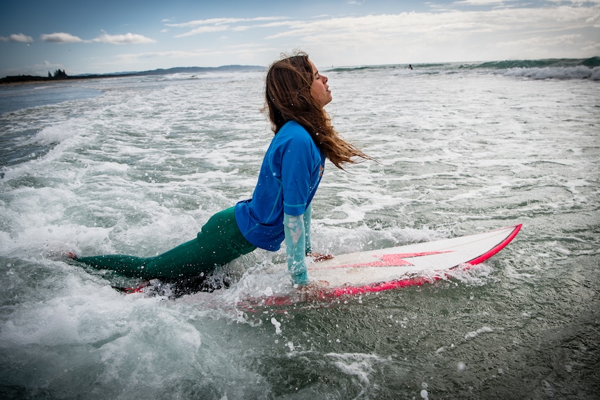 A teenage girl from the side in the ocean jumping on her surfboard as she heads out into the water