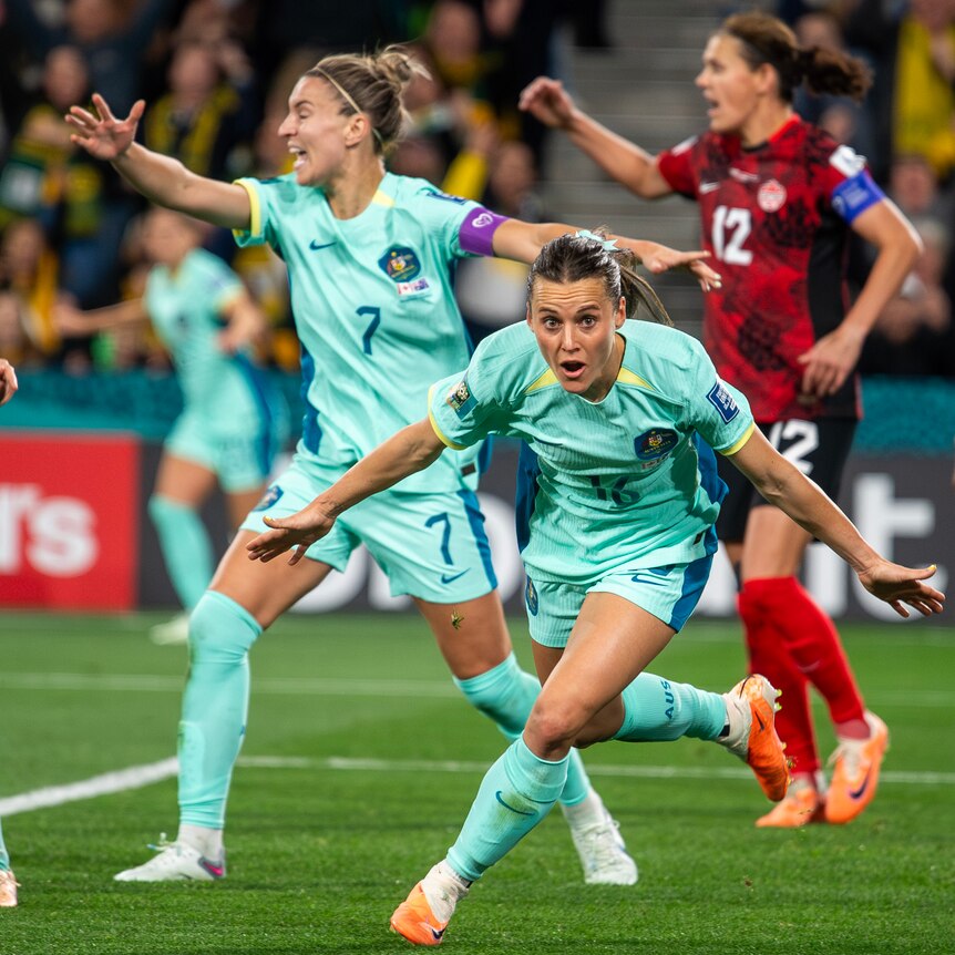 A Matildas striker crouches low to the ground as she runs away from goal with arms spread, as teammates celebrate a goal.