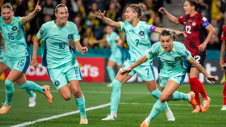 A Matildas striker crouches low to the ground as she runs away from goal with arms spread, as teammates celebrate a goal.
