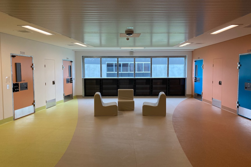 A colourful prison hallway with doors and three chairs in the middle.