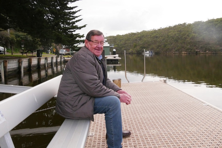 An older man sits on a bench on a river dock. shacks and boats behind him