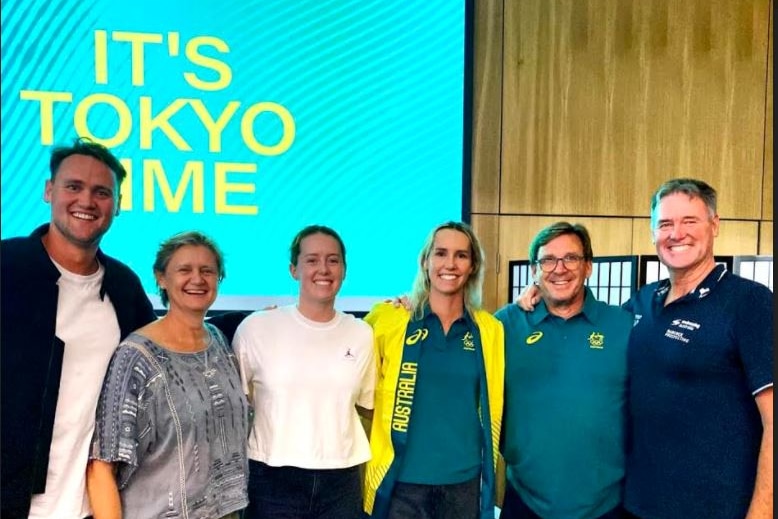 Six smiling people standing arm in arm in front of a sign saying 'It's Tokyo Time'