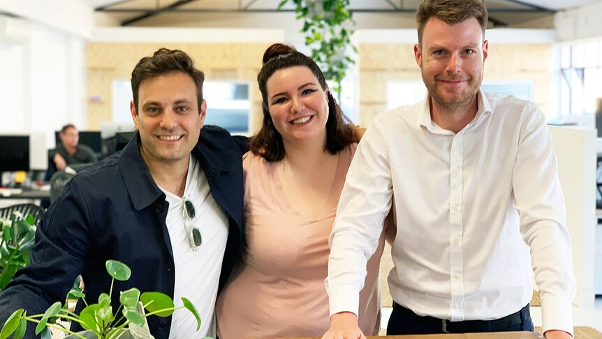 Two men and a woman stand at a timber bench in a modern warehouse beside a pot plant.