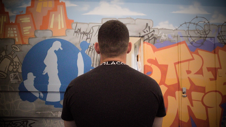 A young man in a black t-shirt sits with his back to the camera, looking at a wall of brightly-coloured graffiti.