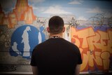 A young man in a black t-shirt sits with his back to the camera, looking at a wall of brightly-coloured graffiti.
