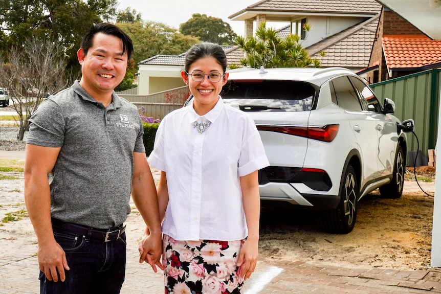 A man in a medium grey polo and his wife, in a white button-down hold hand outside a house with a white electric car behind them
