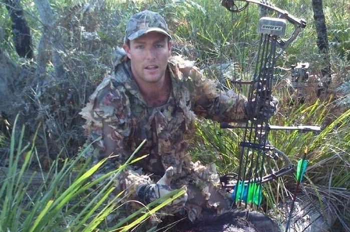 A man crouching outdoors and looking at the camera, with his mouth open.