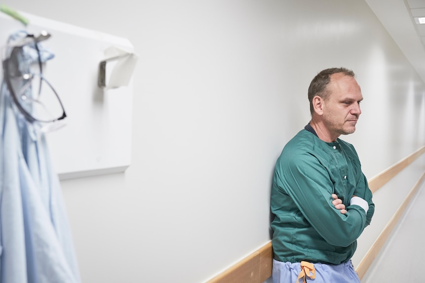 A man wearing a green lab gown stands against the wall in a corridor, next to a blue clinical smock