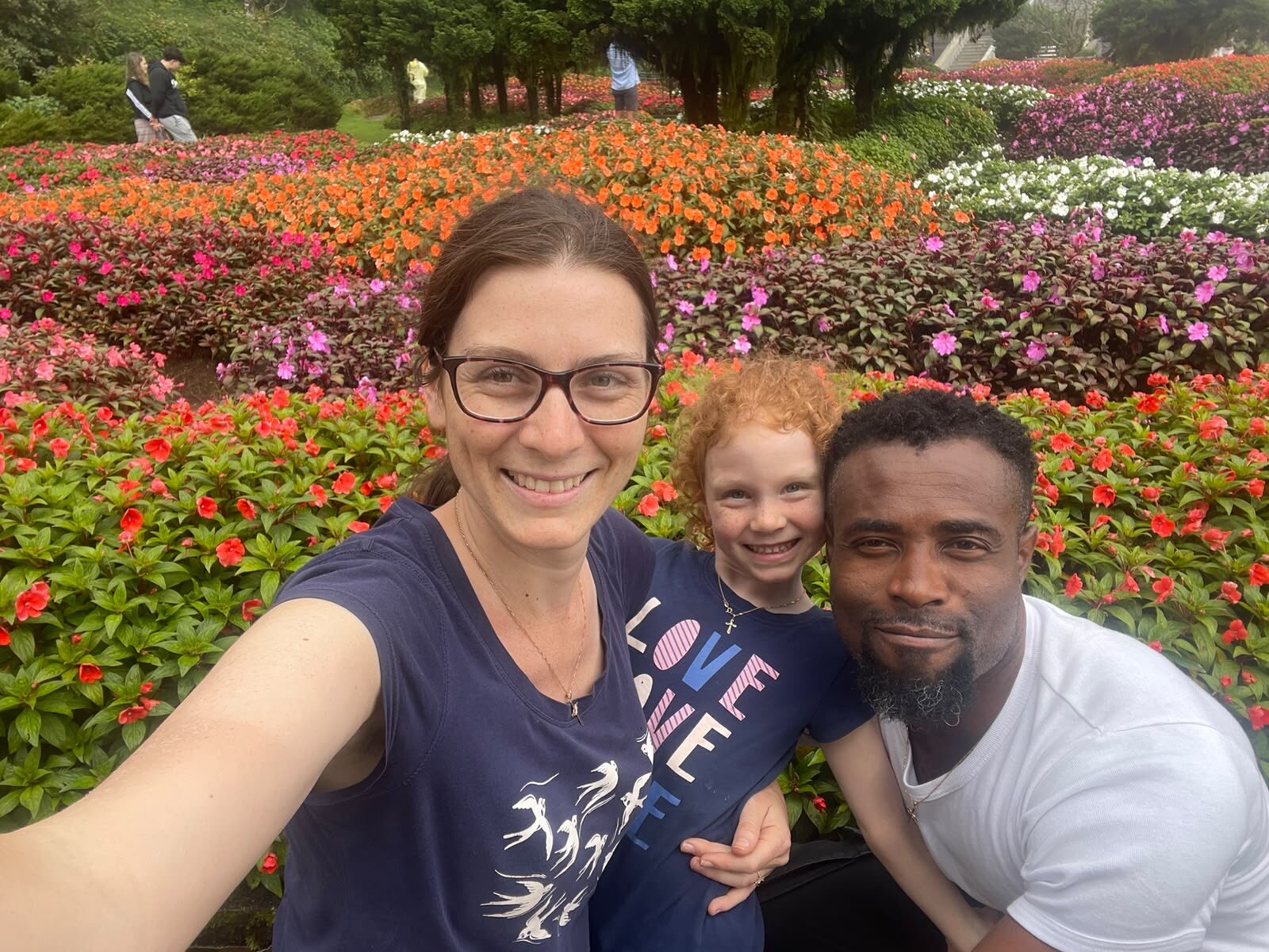 A man and woman with a little girl smilling at camera with flowers in the background.