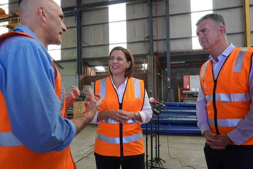 Deb Frecklington and Tim Manger wearing high visibility orange vests and listening to a worker talking