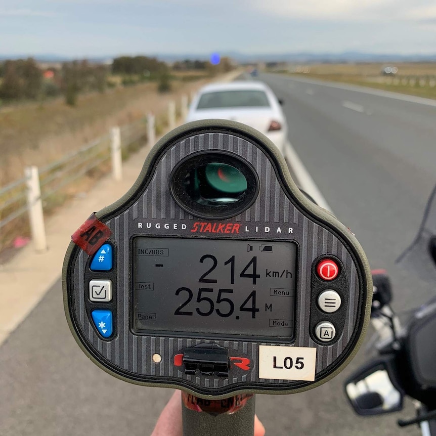 A radar gun shows a detected speed of 214 kilometres per hour. In the background, a car is pulled over on the side of the road.