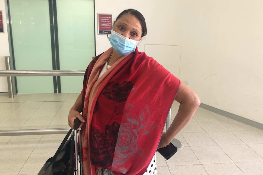 A woman in a facemask and red shawl stands inside an airport terminal carrying a bag and mobile phone.