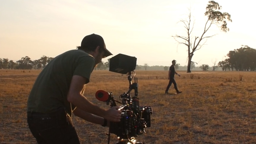 A cameraman films an actor walking through a dusty paddock
