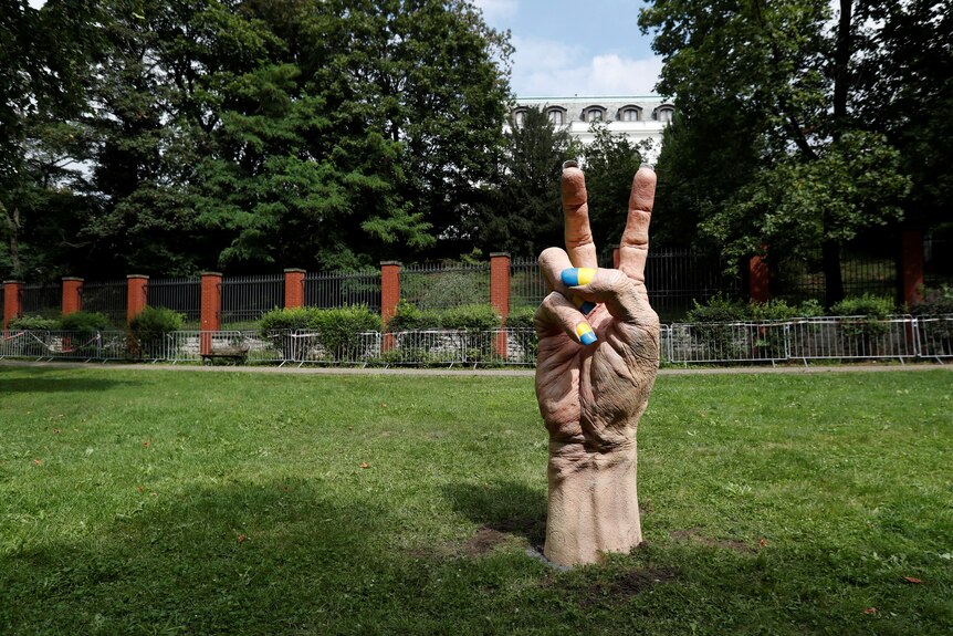 A stone statue of a hand making the peace sign sits on a grassy hillside. Its fingernails are painted yellow and blue.