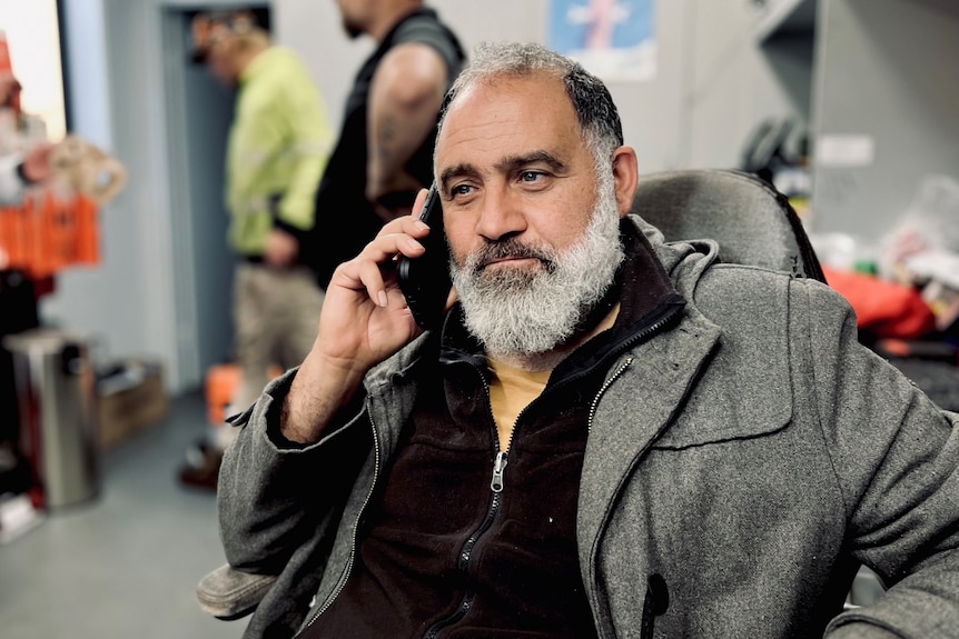 An older man with a greying beard and a grey coat sits in an office chair holding a mobile phone to his right ear.