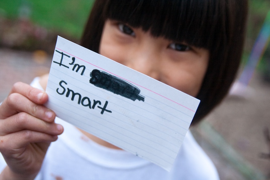 A young girl holding a handwritten note that says 'I'm smart' over most of her face to indicate defeating imposter syndrome