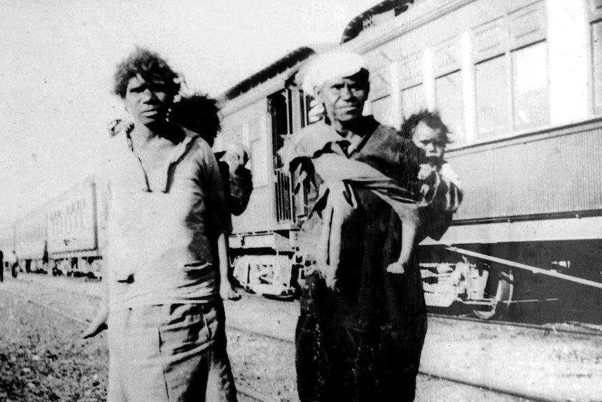 Two women carry children in front of a train at Moore River.