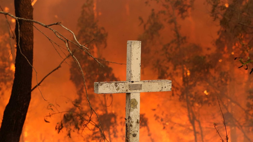 A white cross with chipped paint stands in front of a burning bushfire scene.