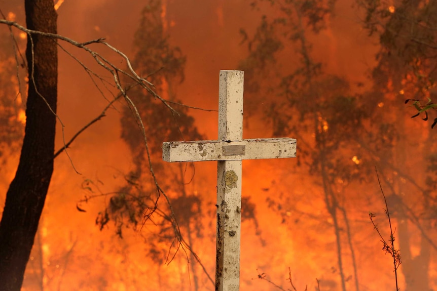 A white cross with chipped paint stands in front of a burning bushfire scene.