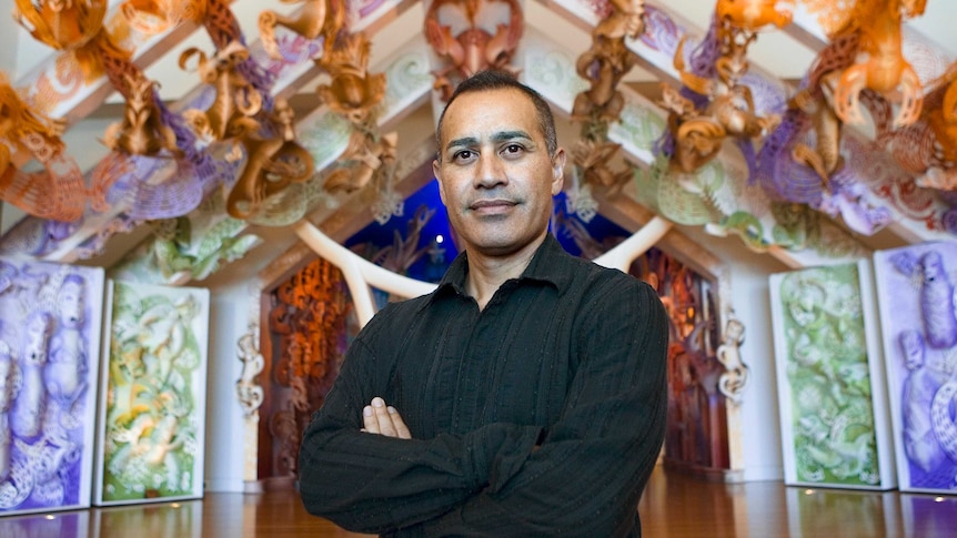 A portrait of a Maori man standing with his arms folded, and a slight smile on his face.