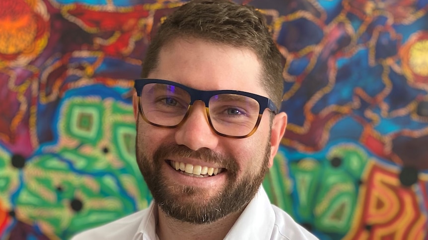 Man smiling at camera wearing glasses and a white shirt, in front of colourful Aboriginal art background