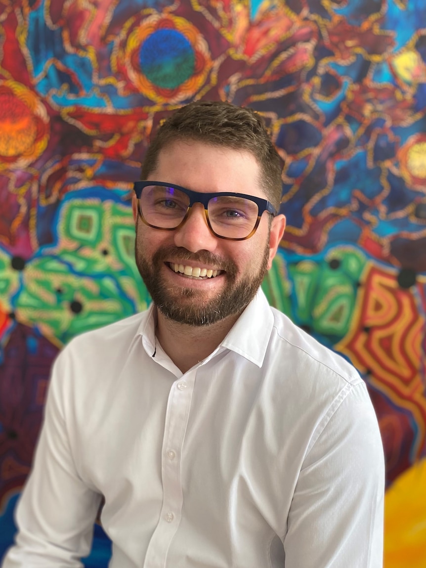 man smiling at camera wearing glasses and a white shirt in front of colorful aboriginal art background