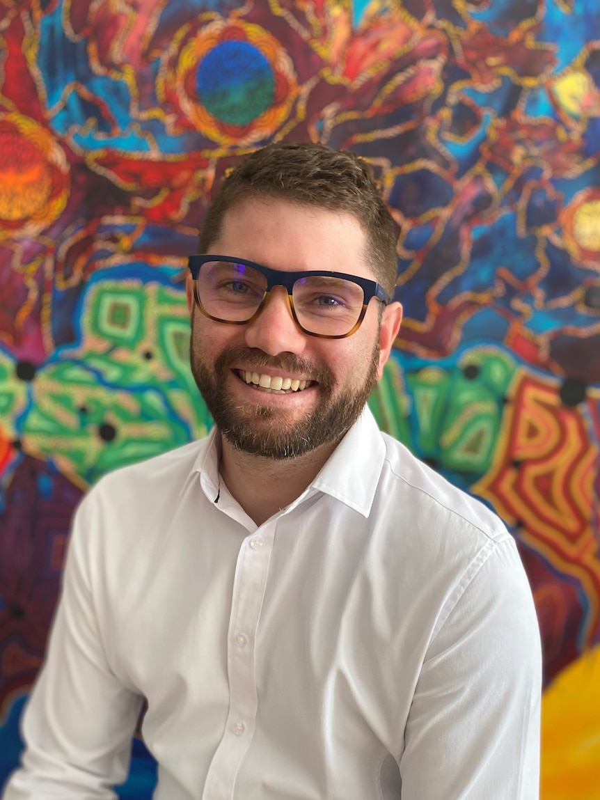 Man smiling at camera wearing glasses and a white shirt, in front of colourful Aboriginal art background