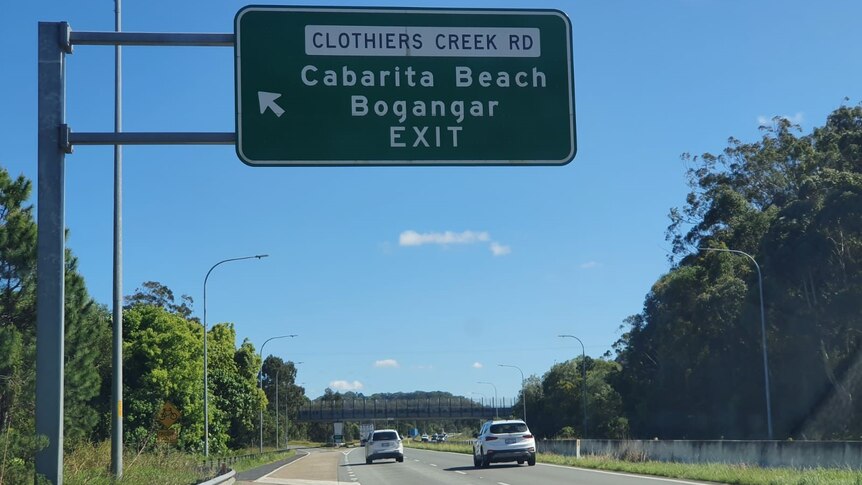 Two cars drive along a motorway under a road sign indicating an exit onto Clothiers Creek Road.