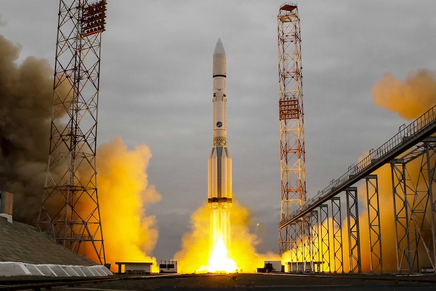 The Proton-M rocket blasts off from the launchpad, with yellow flame coming out of the base of the rocket.