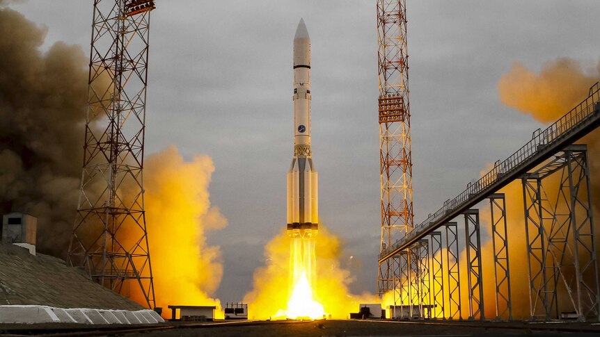 The Proton-M rocket blasts off from the launchpad, with yellow flame coming out of the base of the rocket.