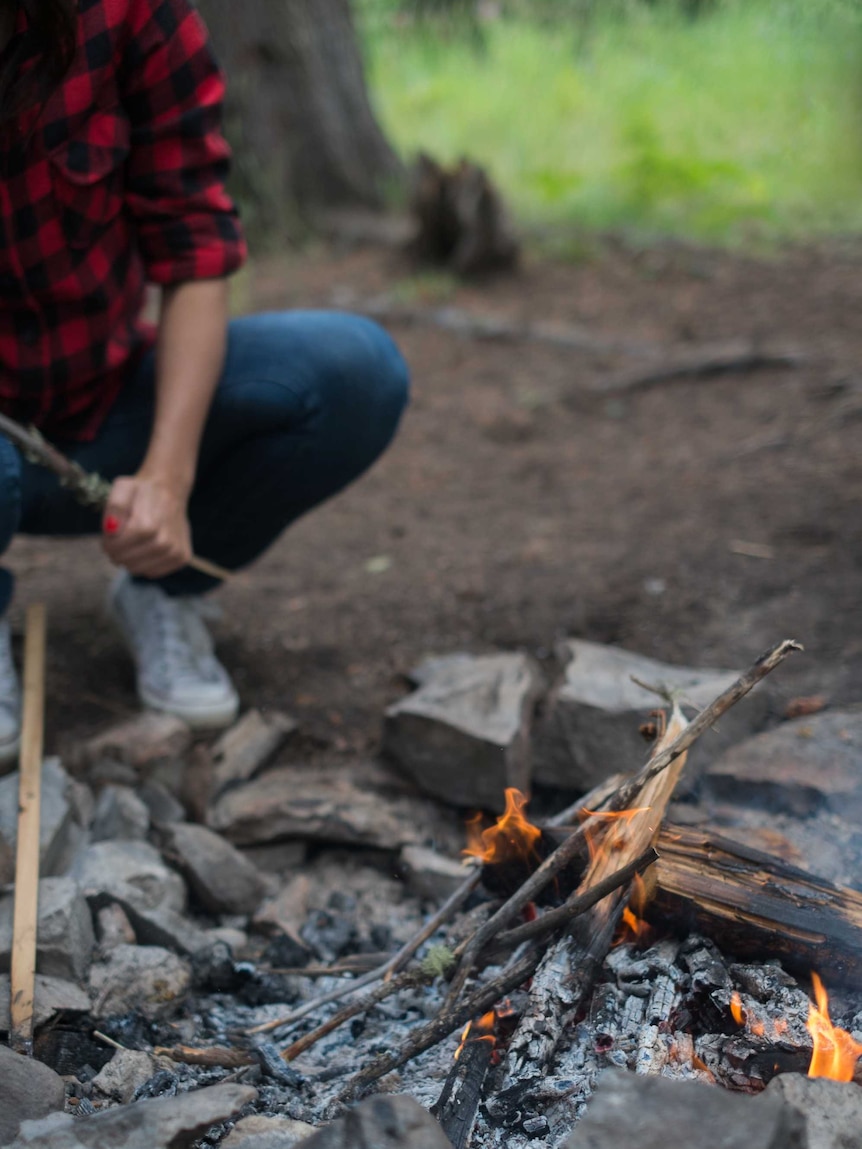 A person squats next to a campfire, which is burning.