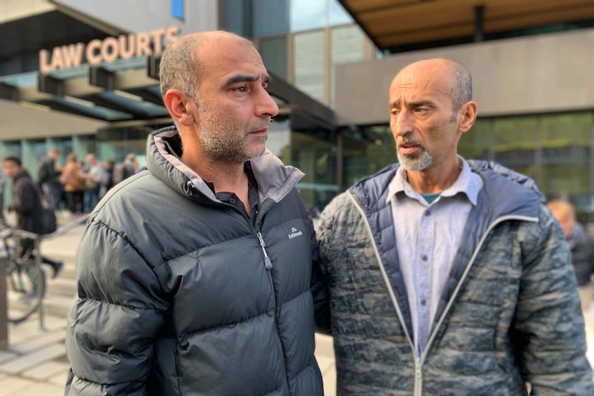 Two tearful men wearing jackets comfort each other while standing outside the Christchurch Law Courts.