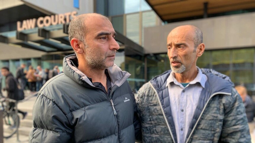 Two tearful men wearing jackets comfort each other while standing outside the Christchurch Law Courts.