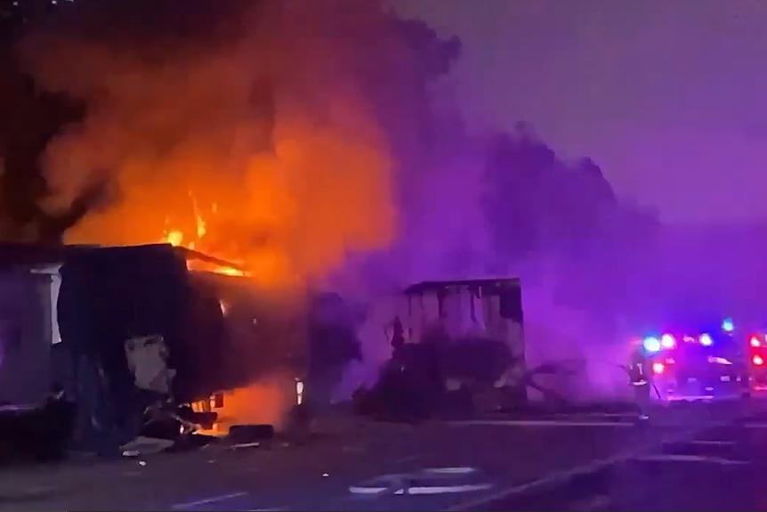 truck engulfed on fire at night, with bright lights of emergency services visible