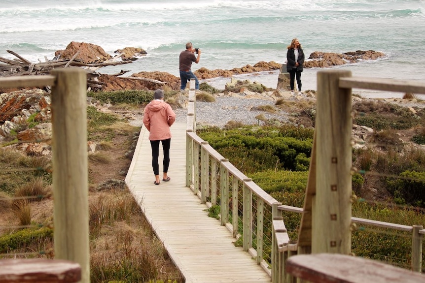 A man takes a photo of a woman near the ocean as a woman walks towards him on a wooden path