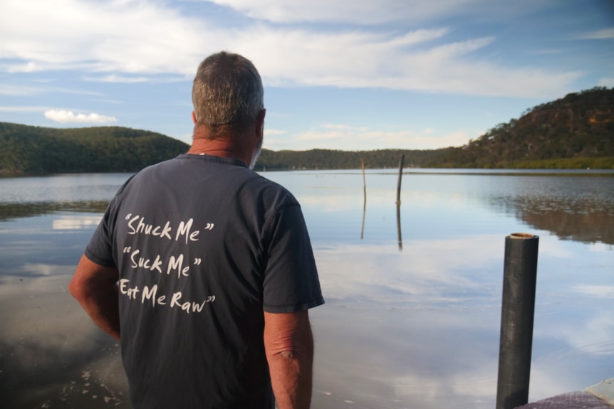 Man looks at river, back of shirt says shuck me, suck me, eat me raw 
