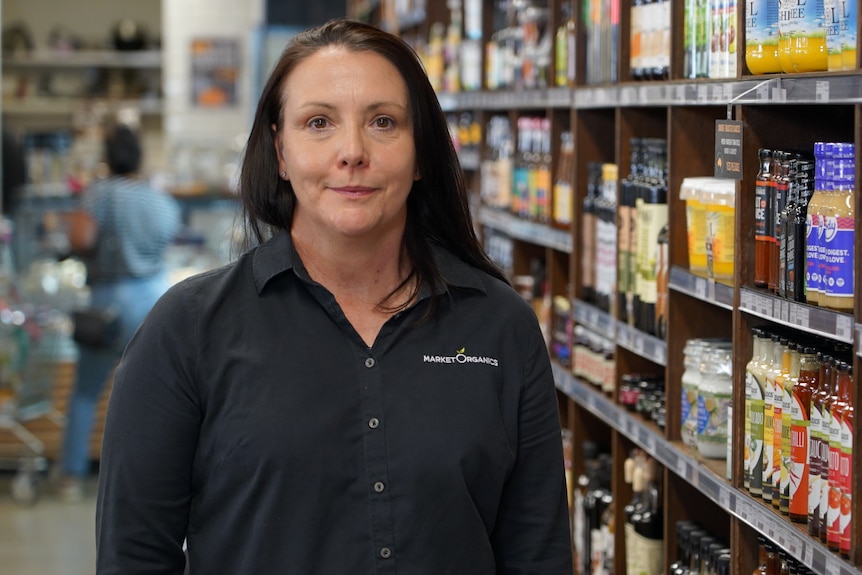 A woman with long, dark hair, wearing a dark shirt, stands in a supermarket.