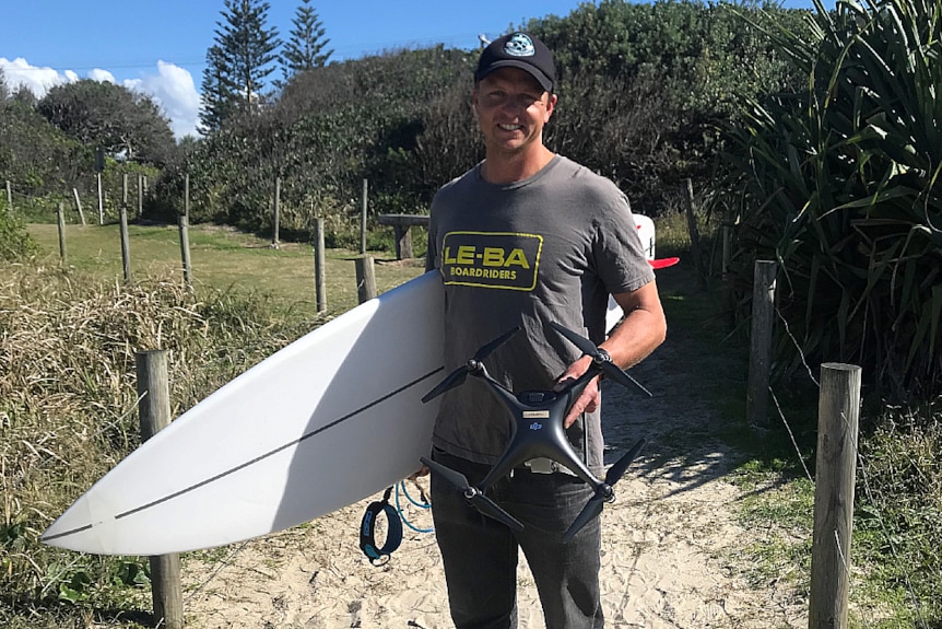 A man is holding a drone in one hand and a surfboard in the other