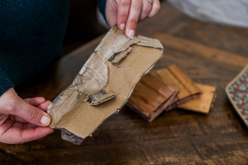 A close-up photograph of a pair of hands holding some cardboard, with wooden floorboards in the background.