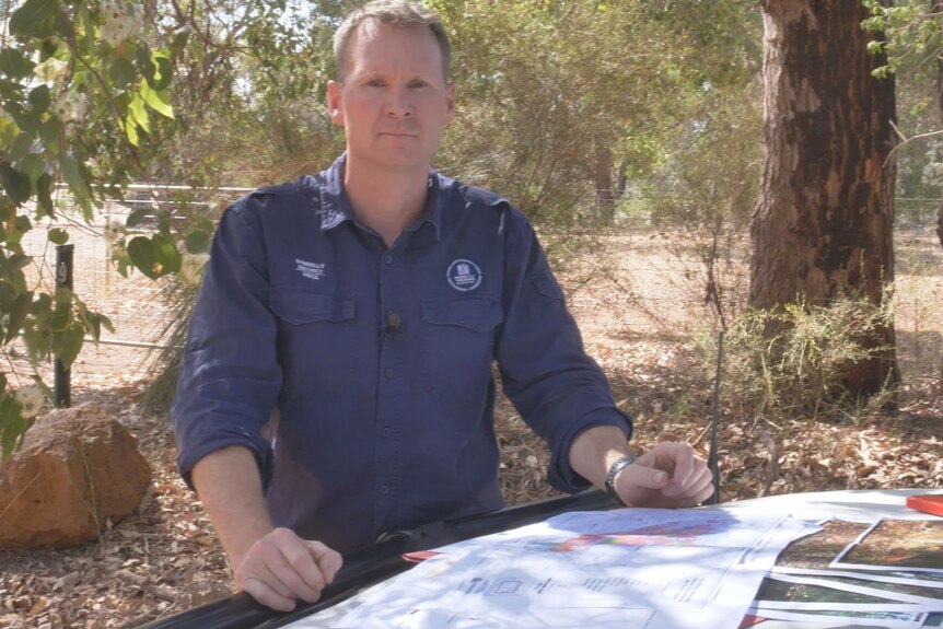 A man with a serious expression standing in the bush, with a map and photos on a table.