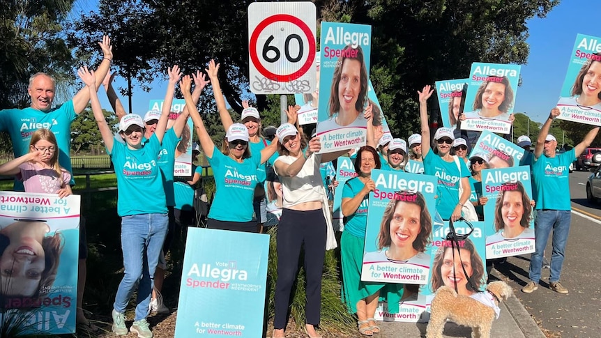 A group of people in teal t-shirts pose for a photo with 'Allegra Spender' posters by the side of a road