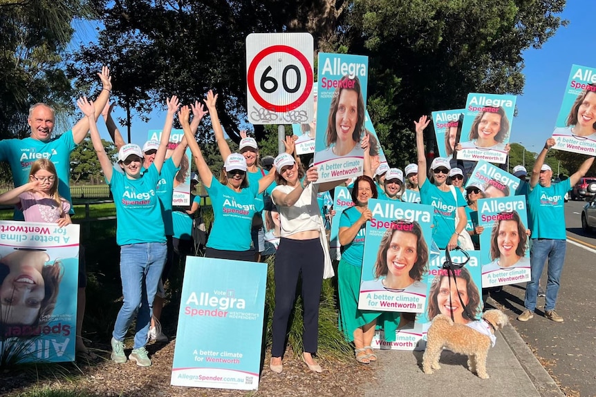 A group of people in teal t-shirts pose for a photo with 'Allegra Spender' posters by the side of a road