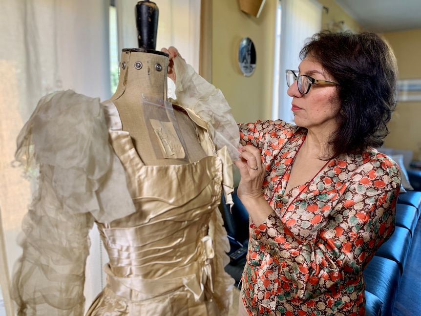 An older woman with dark short hair, wearing glasses, works on a wedding dress on a mannequin.
